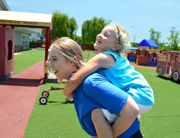 About Merit Schools Childcare - teacher giving child piggyback ride on playground